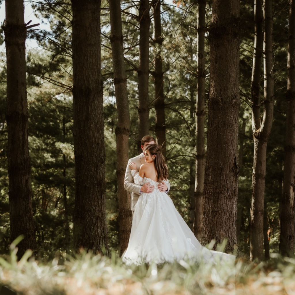 Bride wearing long white dress is turned away from the wedding photographer as she is locked in an embrace with her groom who is nearly entirely obscured from the shot by the bride. Only his arms draped around the bride and one shoulder of his tax tuxedo is visible. The towering evergreens of the enchanted forest at Fort Osage. The thick forest canopy lets in just enough sunlight for a truly fairy tale setting. Now that the outdoor ceremony at Evergreens Chapel near Downtown Kansas City.
