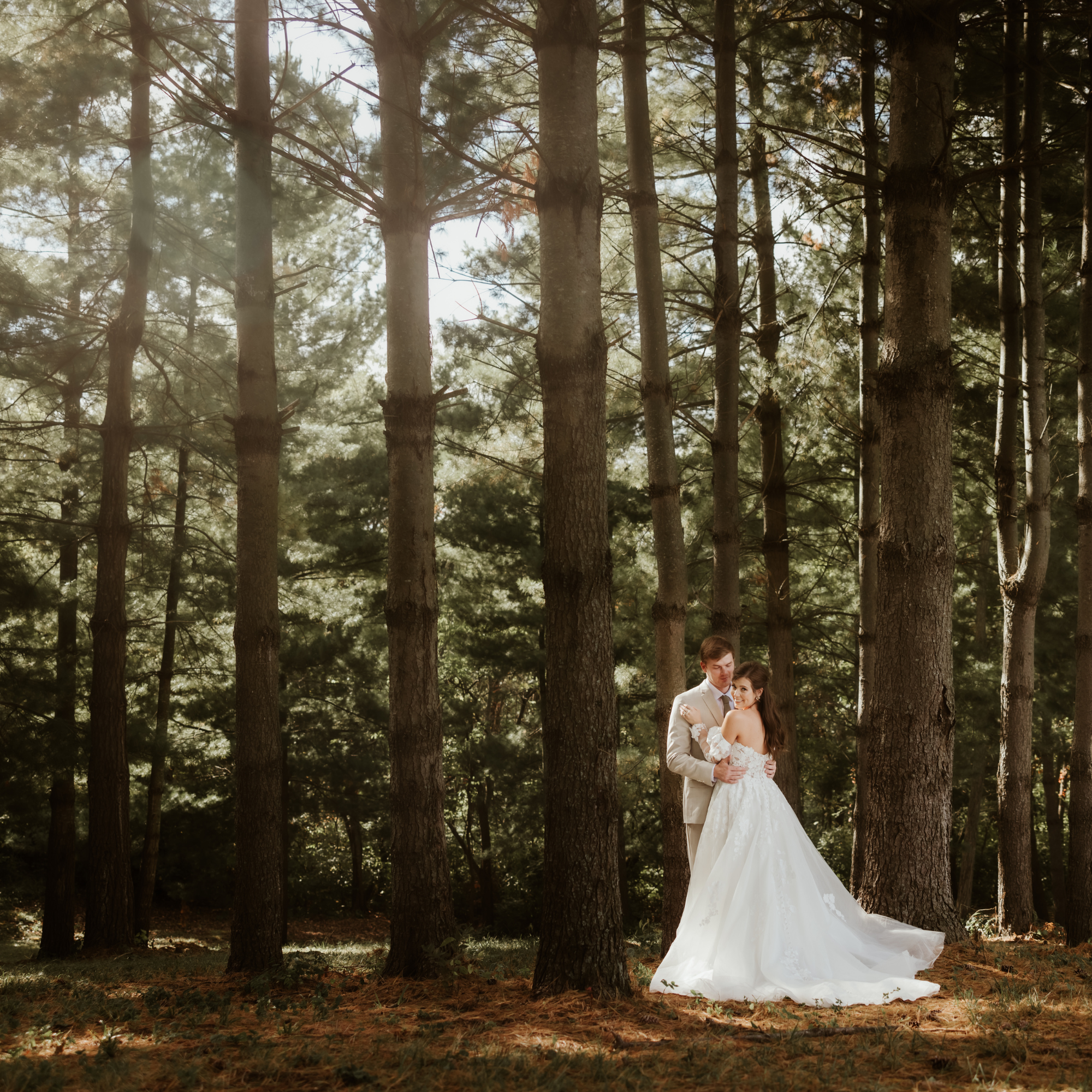 Bride and groom locked in a love embrace in the enchanted evergreen forest wedding venue of Fort Osage Rustic bark and branches of evergreen pine needles filter the sunlight as it shines on the couple and the forest floor of rust orange pine needles. This fairy tale enchanted forest wedding venue is only minutes from Downtown Kansas City at Fort Osage Christmas Trees and Weddings.