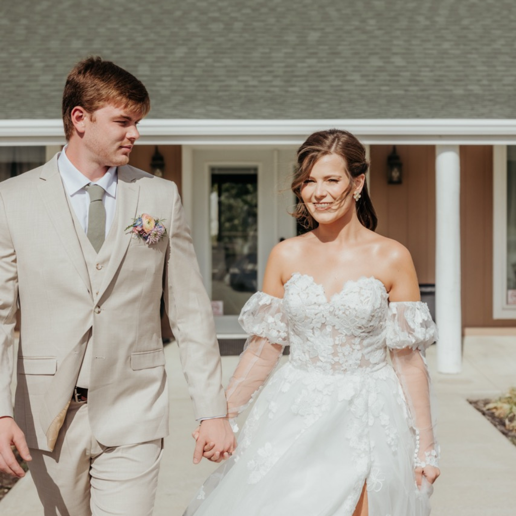Bride wearing white long lace wedding gown holding hand of groom wearing tan tuxedo with green tie and white, peach, blue, and green flowers corsage. The green roof of the White pine lodge fills the back of the picture while the stunning white columns and cement add contrast to the buildings beige paint job.  After the outdoor ceremony at Celestial Glow wedding chapel  the reception was held. Fort Osage Christmas Tree Farm and Wedding Venue near Kansas City is the forest outdoor venue you are looking for.