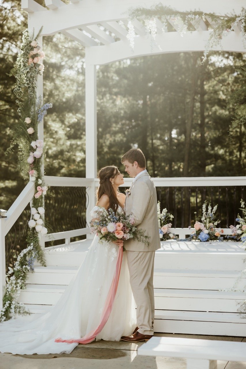 Bride in white dress and groom in tan tuxedo, eyes locked while their faces inch closer for their first married kiss. Behind the couple th the Celestial Glow Chapel is a massive white pergola decorated with peach and blue flowers and greenery. The sun washes out the middle top of the picture as it shine through the enchanted forest canopy in the background. This fairy tale enchanted forest setting of Celestial Glow Chapel of Fort Osage Christmas Tree Farm and Wedding Venue Kansas City