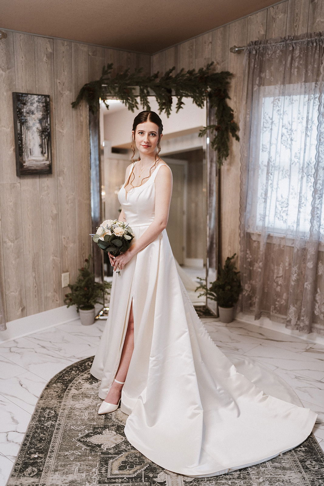 Woman in a white wedding dress holding flowers in an evergreen Christmas tree farm decorated room.