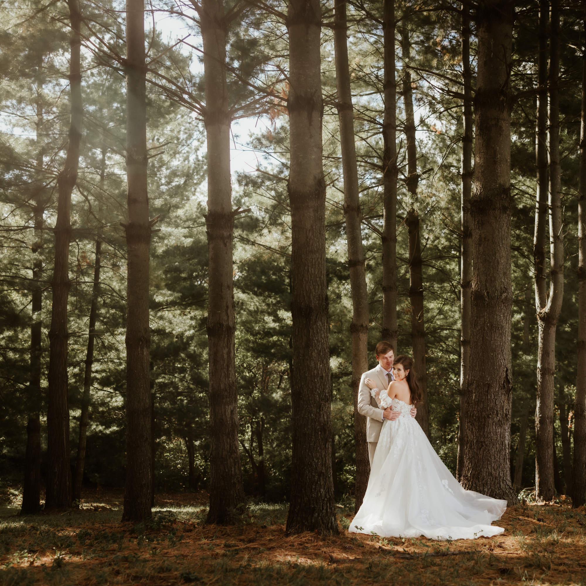 Bride and groom locked in a love embrace in the enchanted evergreen forest wedding venue of Fort Osage Rustic bark and branches of evergreen pine needles filter the sunlight as it shines on the couple and the forest floor of rust orange pine needles. This fairy tale enchanted forest wedding venue is only minutes from Downtown Kansas City at Fort Osage Christmas Trees and Weddings. 