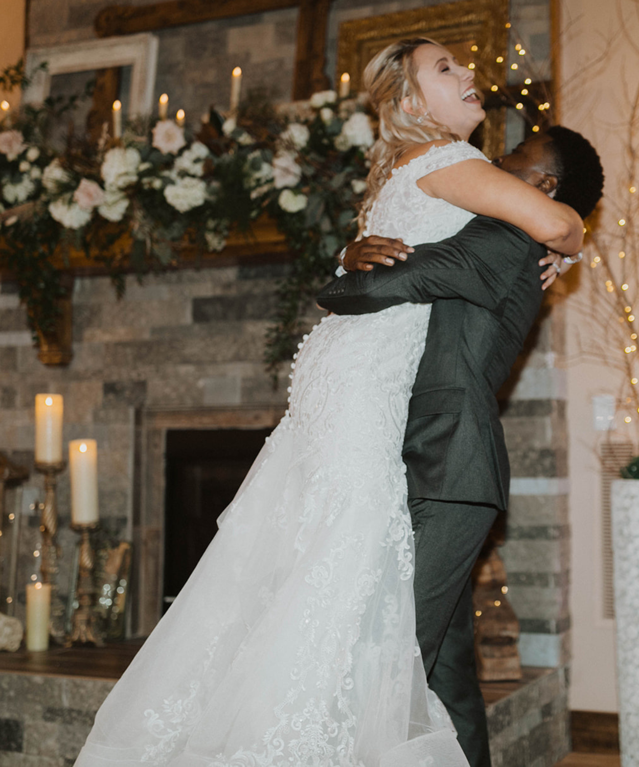 Groom lifts up bride during spirited first dance on the ballroom at White Pine Lodge.  Her long white gown is stunning while her laughter fills the room.  Behind them a beautiful fireplace decorated with decorative frames white flowers candles and gold accents. This wedding reception at White Pine Lodge at Fort Osage Christmas Tree Farm and Wedding venue in Kansas City