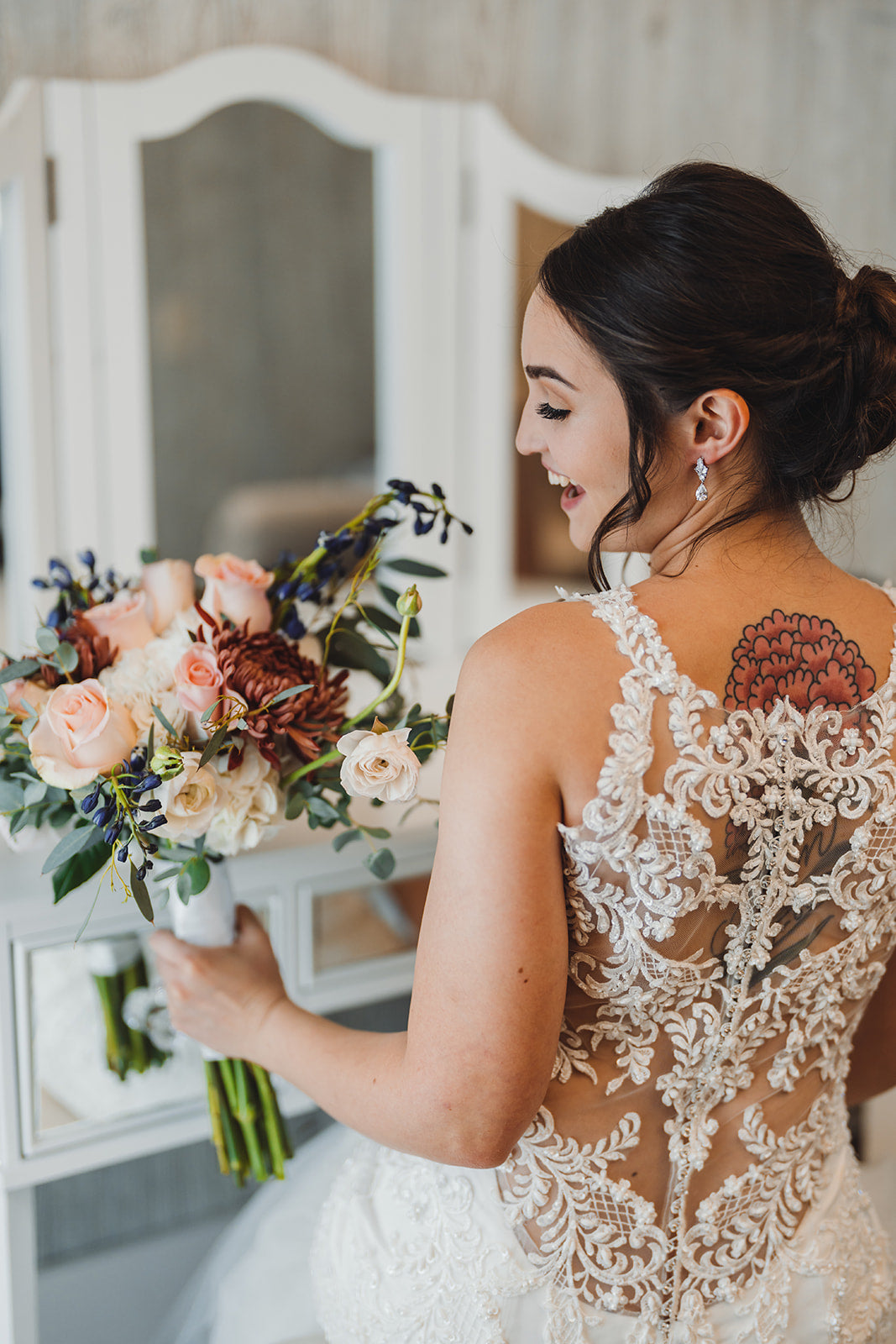 Glowing bride holding her bouquet of lush flowers of peach, burgundy and blue at the fairy tale mirrored vanity of the bridal suite at White Pine Lodge. The back of her intricately laced wedding dress exposing a floral tattoo that matches perfect the burgundy flower in her bouquet. Her smile is radiant as she prepares in the comfort of the bridal suite and anticipation builds for the outdoor ceremony at Celestial Glow Chapel of Fort Osage Christmas Tree Farm and Wedding Venue of Kansas City