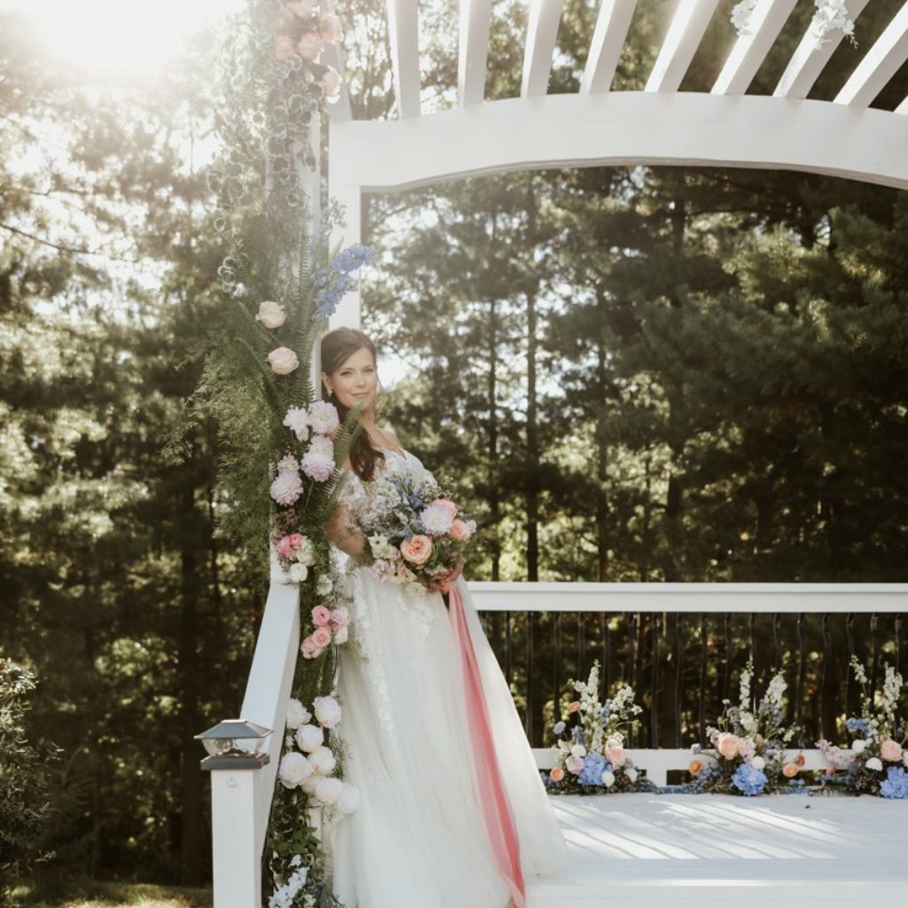 Bride standing in the pergola of Celestial Glow Chapel in the enchanted evergreen forest of Kansas city during her outdoor wedding ceremony. She is wearing a long white wedding gown and smiling as she holds her bursting bouquet of white, peach, and blue flowers with green accents. More flowers with long branches of green fern are woven around the pergola. A celestial glow is seeping through the majestic forest canopy overhead.  Celestial Glow Chapel at Fort Osage Christmas Tree Farm and Wedding venue