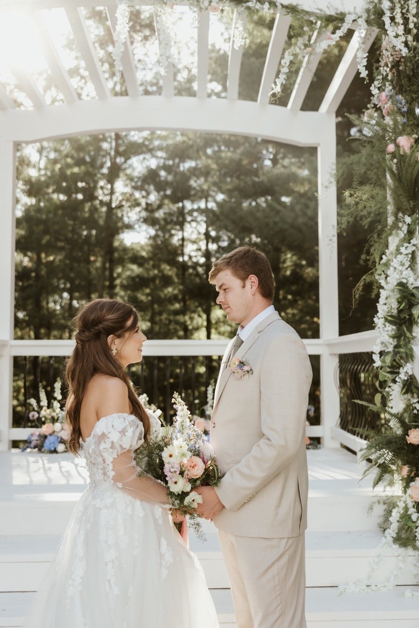 Bride and groom hold hands under the pergola of Celestial Glow Chapel in the Enchanted Evergreen Forest of Kansas City. Her White dress with delicate lace flowers and his tan tuxedo are illuminated with a celestial glow filtering through the evergreen canopy. The forest trees are the perfect backdrop for their outdoor wedding ceremony at Fort Osage Christmas  Tree Farm and Wedding Venue