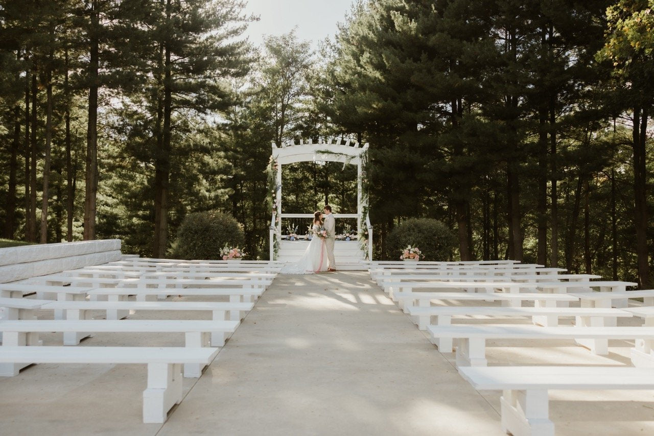 A bride and groom embrace at Evergreens Chapel at Fort Osage venue. The outdoor wedding ceremony is surrounded by enchanted evergreen forest. Tall majestic pine trees tower over the chapel and the sun dramatically shines through the bark covered tree trunks and branches filled with evergreen pine needles. The contrasting white paint of the large pergola and benches against the giant pines is breathtaking. It impossible to believe that this fairy tale forest wedding venue is just minutes from Kansas City