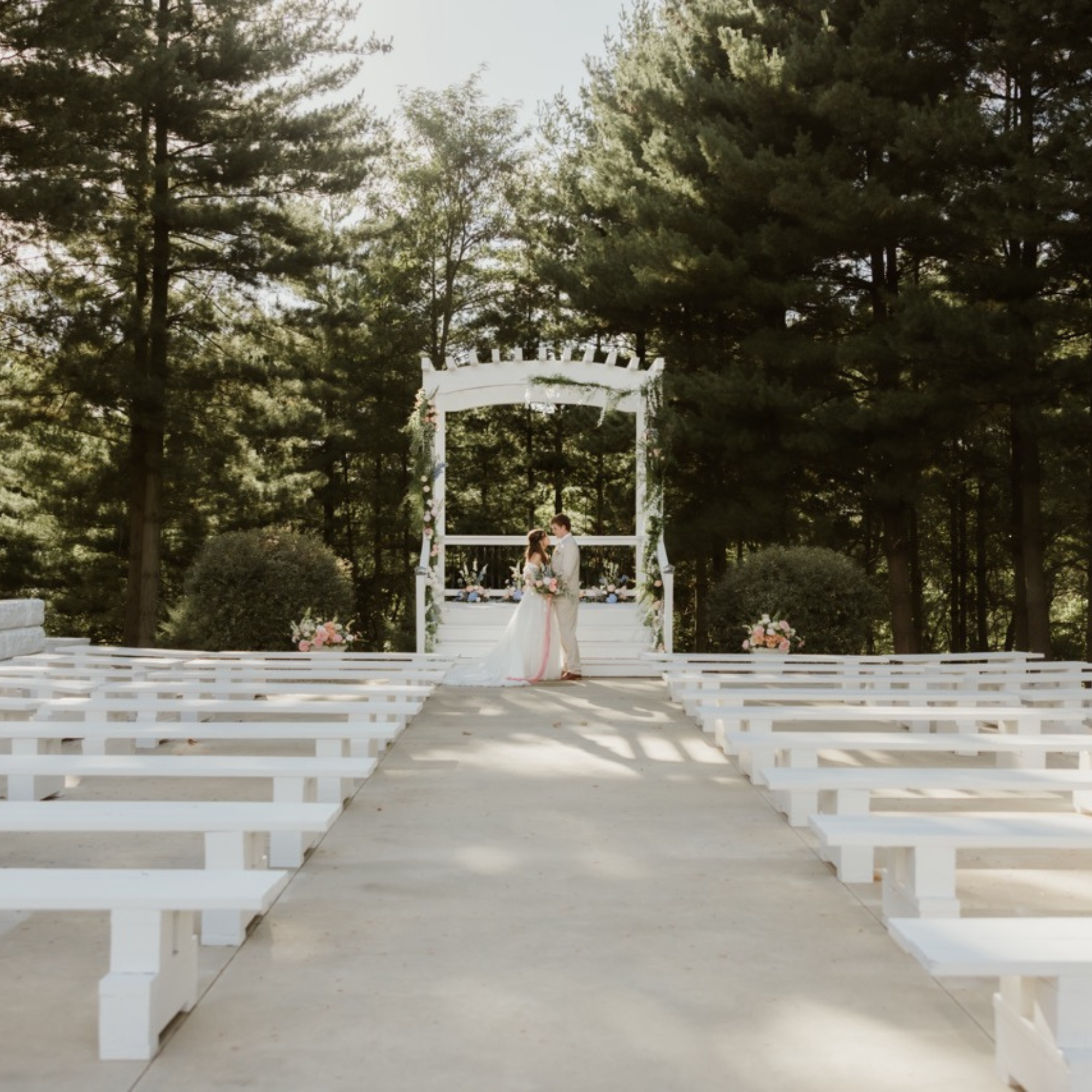 A bride and groom embrace at Celestial Glow Chapel in the enchanted evergreen forest of Kansas City. The outdoor ceremony space is surrounded by tall majestic pine trees towering overhead to create a cathedral of nature. The sun dramatically shines through the bark covered tree trunks & branches filled with evergreen pine needles backing the scene in a Celestial glow. The white paint of the large pergola and benches against the pines is breathtaking. Fort Osage Christmas Tree Farm and Wedding venue in KC