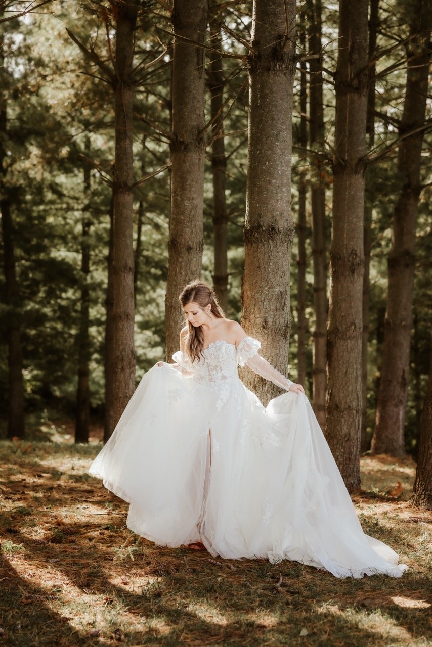 Bride with long brown hair holding up flowing white wedding dress as walks across the enchanted evergreen forest of Kansas City forest floor. Pine needles and celestial light cover the forest floor. Towering evergreens covered rich green pine needles. The bride moves to a new setting during her outdoor wedding ceremony at celestial glow chapel Fort Osage Christmas Tree Farm and Wedding Venue