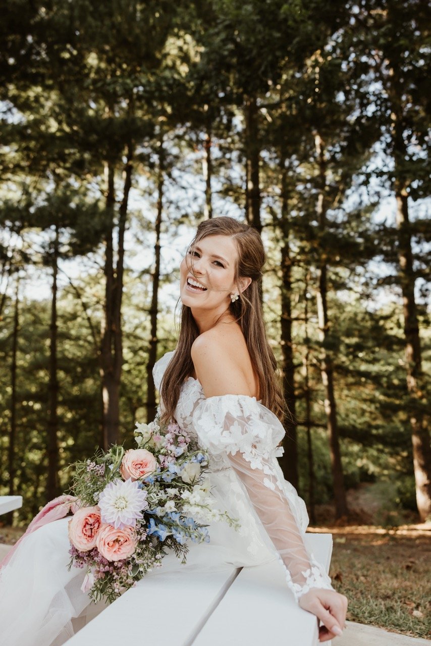 Bride smiling as she holds her wedding bouquet of pink, white, and blue flowers. Her white dress with lace sleeves stands in contrast to the enchanted evergreen forest wedding venue towers overhead. The sun and sky is barely visible through the trunks and pine covered branches of the majestic seventy foot tall white pines of Evergreen's Outdoor Wedding Chapel. The enchanted evergreen forest of Fort Osage Christmas Trees & Weddings is just mins drive away from downtown Kansas City in Independence, Missouri