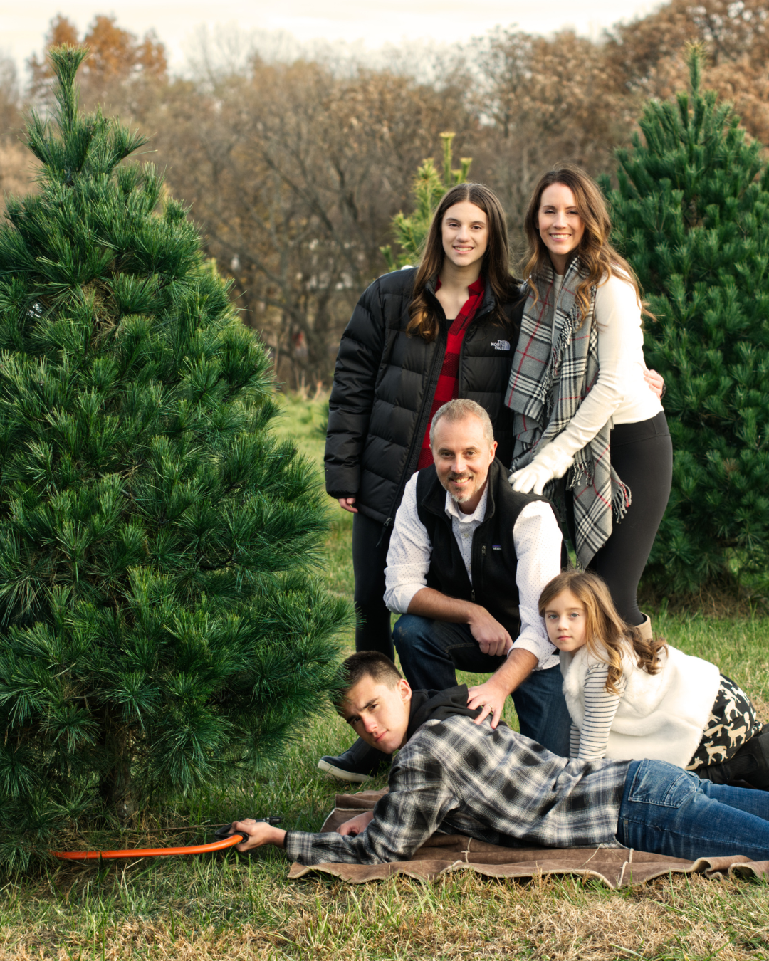 In Kansas City a family of five poses while cutting down a real Christmas tree from Fort Osage Christmas trees and wedding venue. Family is wearing black and white solid and plaid coordinates. The green forest of trees around them are almost as bright as the red buffalo check shirt one of the kids is wearing under their black jacket. Fort Osage Christmas Tree Farm is mins from Downtown Kansas City!