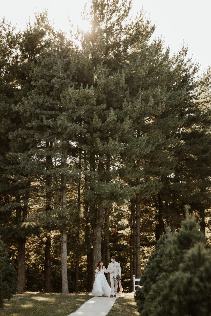 A newly wed couple walks away from the Evergreens Chapel during thier outdoor wedding ceremony at Fort Osage Christmas Trees & Wedding venue with their arms locked together. Massive 70 ft white pine evergreen trees tower above the pair. Sun is shining through the tops of the trees giving the whole scene a hue of heavenly gold. Hard to believe this mountain forest wedding venue is just mins from downtown Kansas City