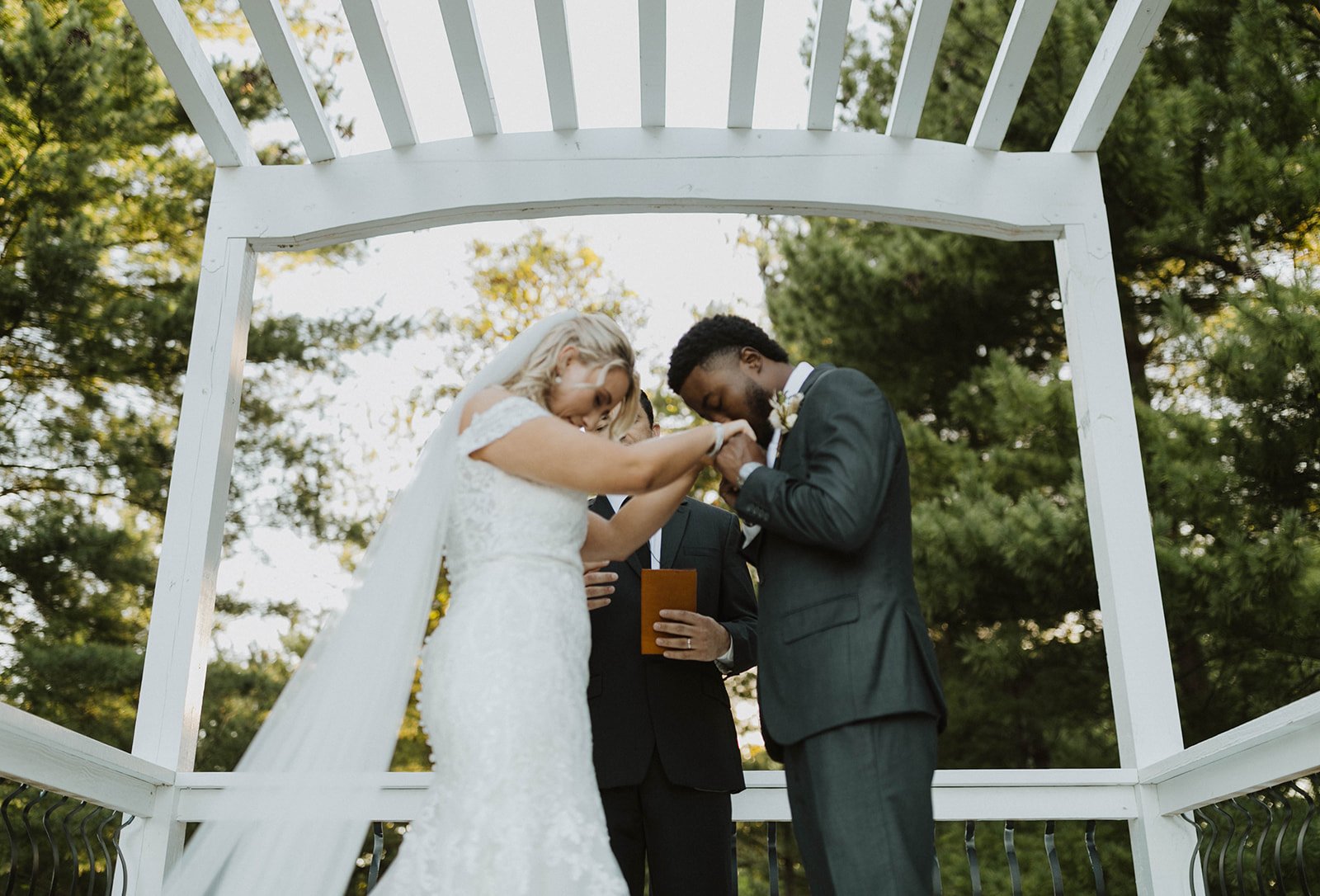 During Outdoor Ceremony a bride wearing white dress holding the hands of her groom as he kisses them and they bow their heads during the outdoor wedding ceremony beneath a white pergola and towering evergreen forest on all sides and sun light filter through. This enchanted forest wedding at Fort Osage Christmas Trees and Wedding venue is only mins from downtown Kansas city