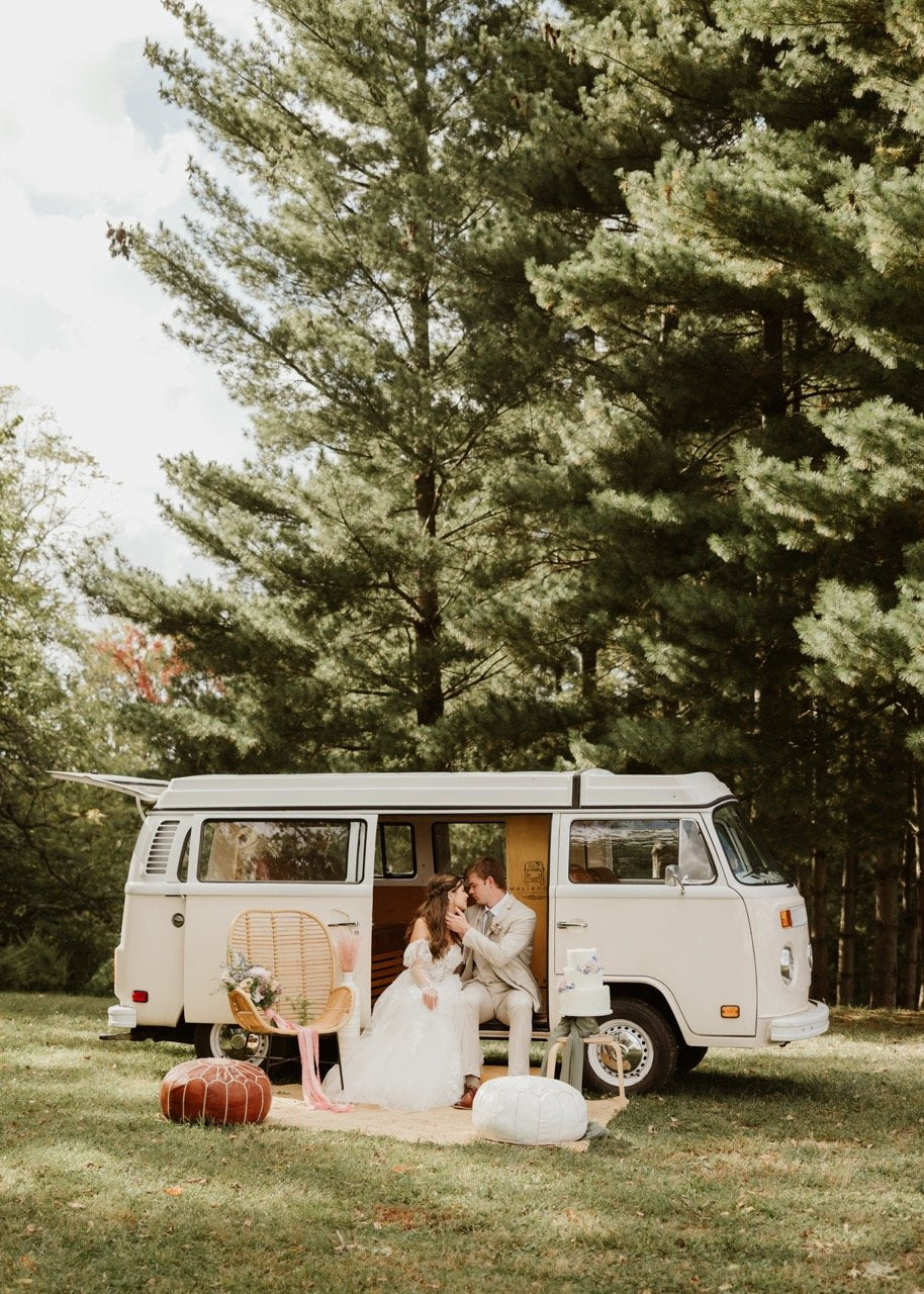 In an Enchanted Forest wedding venue a groom in tan tuxedo places hand on the neck of his bride wearing a long white wedding dress as he moves in for a kiss. They are sitting in the open sliding dour of a vintage volkswagon bus that is beige and white just after the Outdoor ceremony ended at celestial glow chapel. bus is parked in the grass and towering 50 ft evergreen trees are seen in the background. Cant believe that Fort Osage Christmas Tree Farm and Wedding Venue