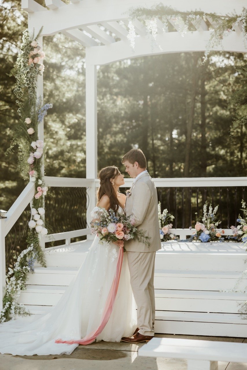 Bride & groom embracing under the pergola at Evergreen's Chapel of Fort Osage during their Outdoor Wedding Ceremony. Her white dress and his tan tuxedo stand in contrast to the towering evergreen enchanted forest behind them. The sun cascades through the canopy kissing the delicate white, peach, and blue blossoms in the brides bouquet and the floral arraignments adorning the pergola. It is a picture perfect outdoor wedding ceremony at The beautiful Fort Osage Christmas Tree and Wedding Venue of Kansas City