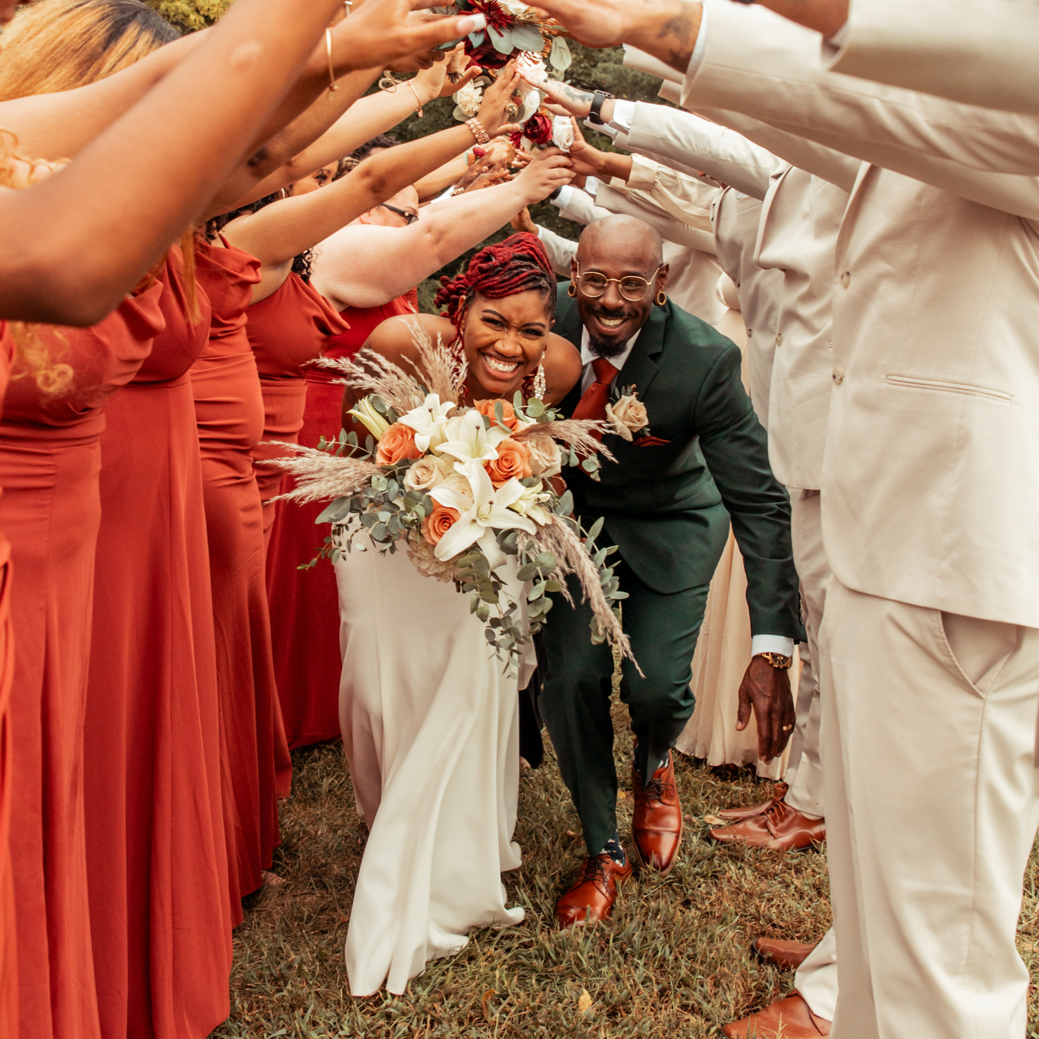 Newly wed Couple celebrating their marriage during outdoor ceremony by running beneath the arms of their wedding party like a tunnel. The bridesmaids are wearing dresses in a rust orange red shade and the groomsmen are wearing ivory tuxedos. The smiling bride is holding a bouquet of flowers so lush they look almost as if they are exploding like a firework of light orange, white Lilly, and green and gold accents. Enchanted Forest Venue at Fort Osage Christmas mins away from downtown Kansas City Missouri.