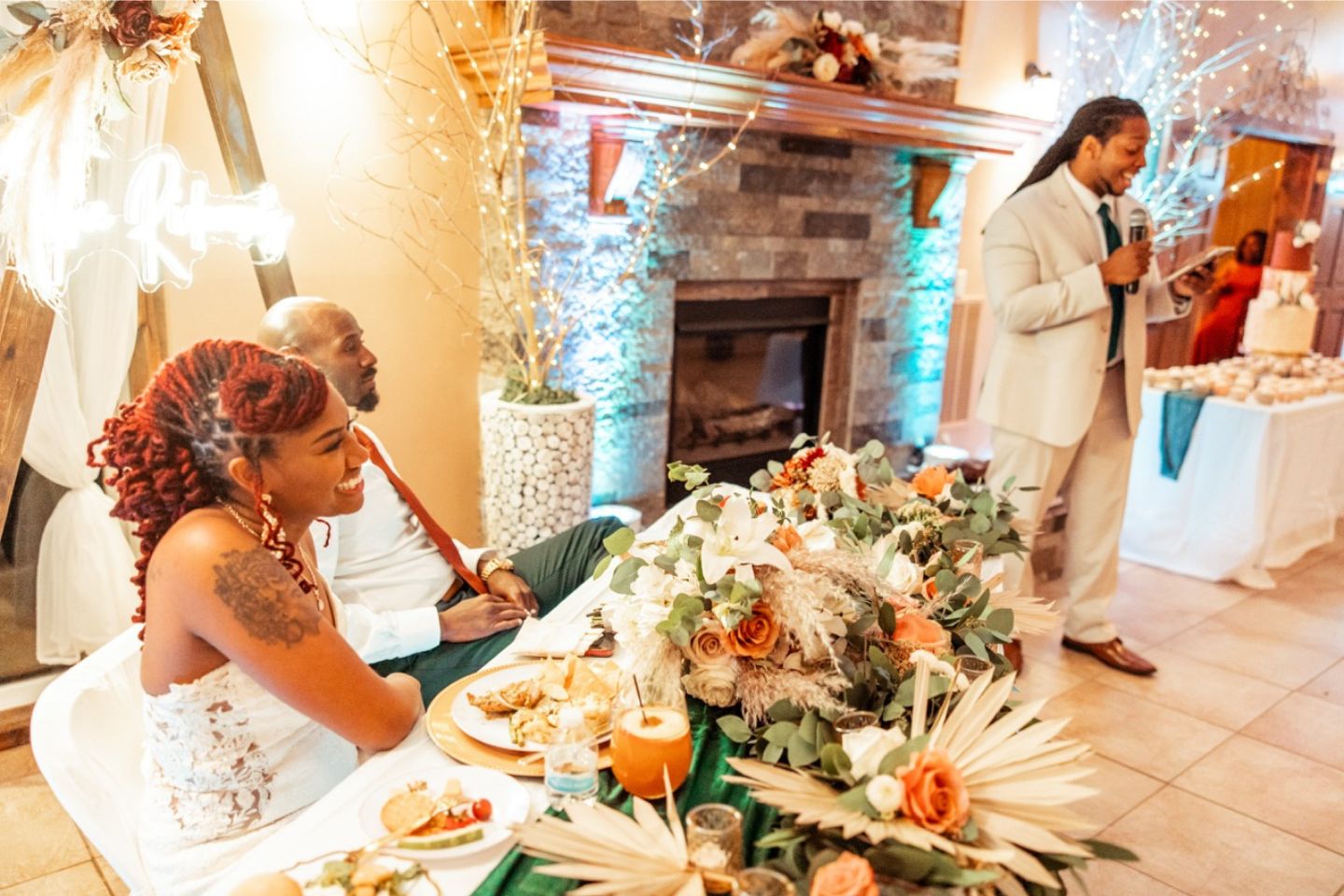 Bride and groom sitting at head table during reception. Warm light fills the picture from enchanting twinkle lights and a glowing sign behind the couple. A groomsman is toasting the couple as he reads prepared remarks he is holding into the microphone. The wedding cake is faintly visible in the back of the photo. Beautiful Wedding Reception at Fort Osage Christmas Tree Farm and Wedding Venue in Kansas City