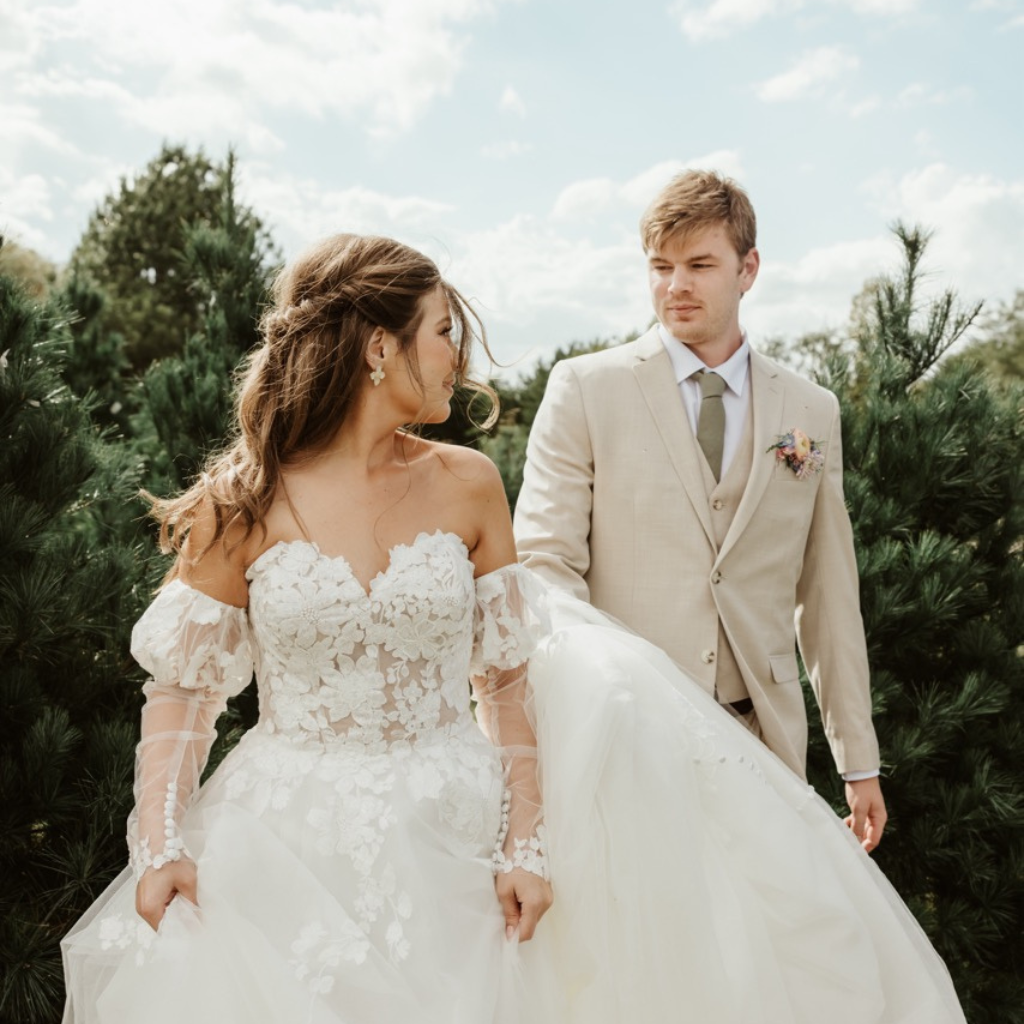 Woman wearing floral lace white wedding gown walks through Christmas tree farm as she looks back at her trailing groom wearing tan tuxedo and vest with sage green tie. Couple is walking in bliss away from their outdoor wedding ceremony at celestial glow chapel at the enchanted evergreen forest of Kansas City at Fort Osage Christmas Tree Farm and Wedding Venue