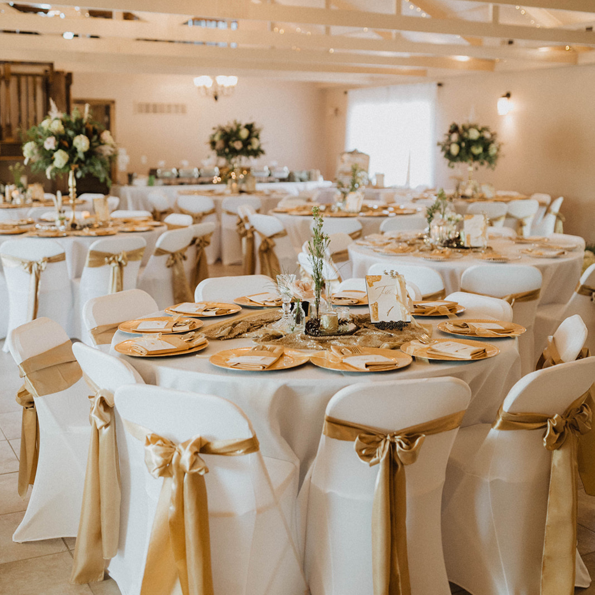 White Pine Lodge at Fort Osage Wedding Venue ready to host a reception in the fall. A half dozen large round tables with white linens, gold place settings, and green accents fill the room. The beige walls are awash in soft light from the wall sconces and sunlight flowing through the windows. The air is filled with anticipation as guest are expected to arrive soon. Located in Kansas City