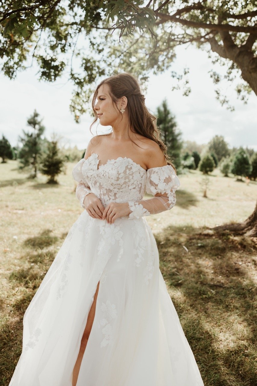 Women with long brunette hair mostly pulled back except for several strands flowing along her face looks on as she wears a long white lace flowing wedding dress on a enchanted Christmas tree farm. She is preparing for her outdoor wedding ceremony at Celestial Glow Chapel at The enchanted evergreen forest of Kansas City Fort Osage Christmas tree farm and wedding venue.